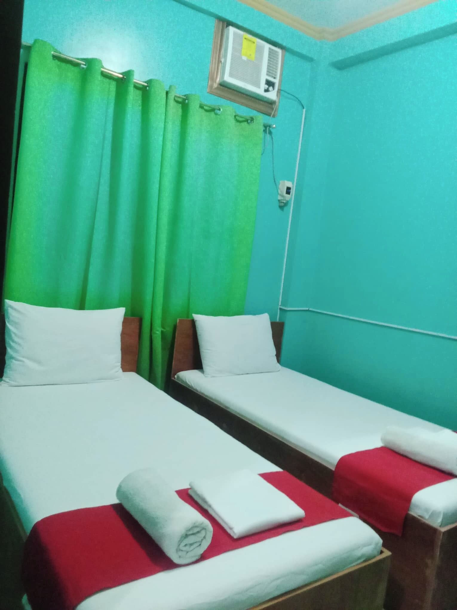 A room with two single beds, green curtains, a wall-mounted air conditioner, and a red and white bedspread.