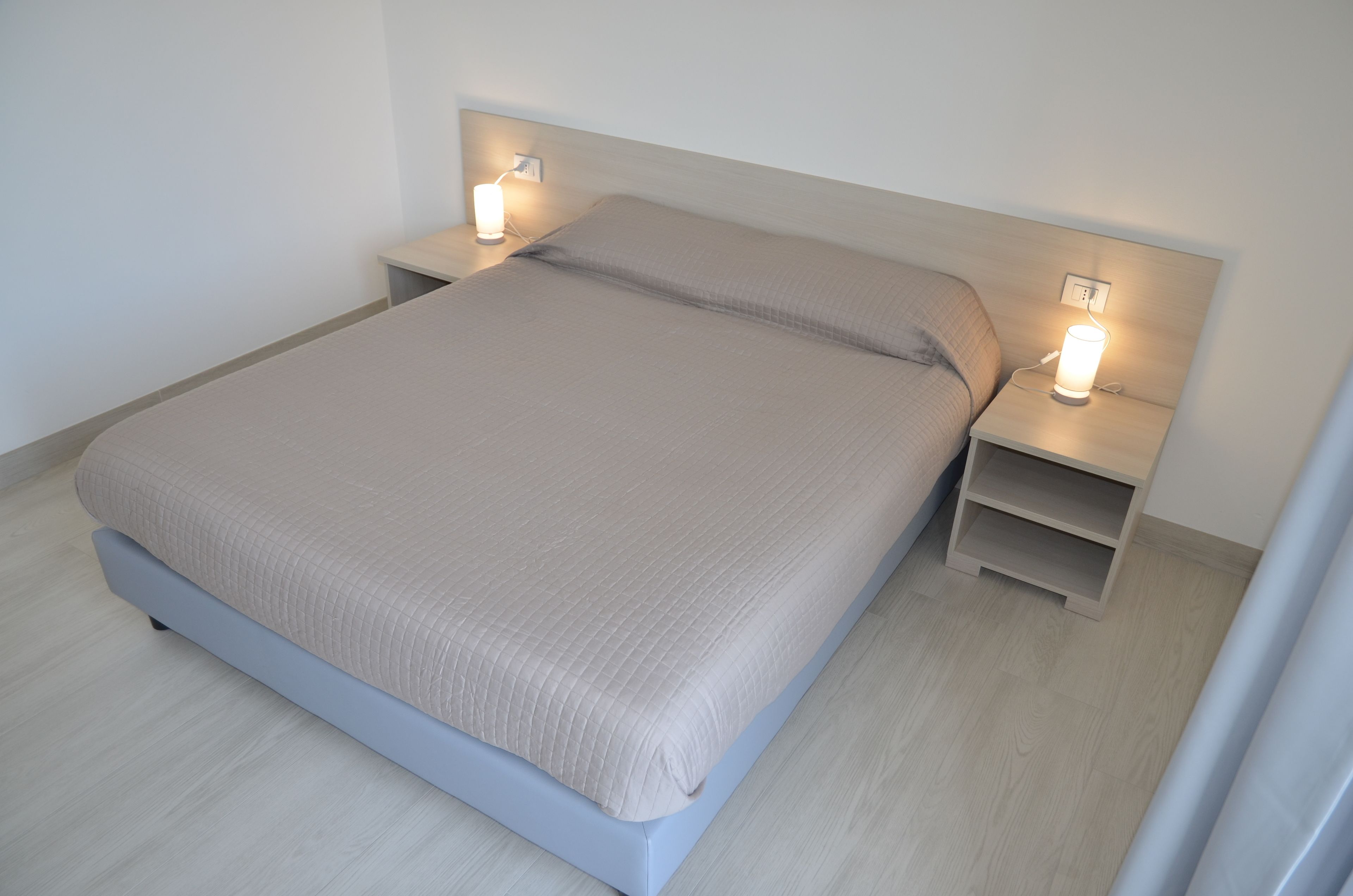 Een net opgemaakt bed met een lichtkleurige dekbedovertrek en kussens, geflankeerd door twee nachtkastjes met lampen, in een kamer met licht houten vloeren en een effen witte muur.