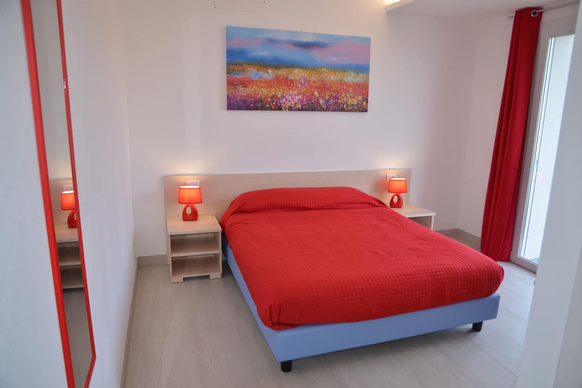 A bedroom with a bed, two bedside tables with lamps, a painting on the wall, and a window with curtains.