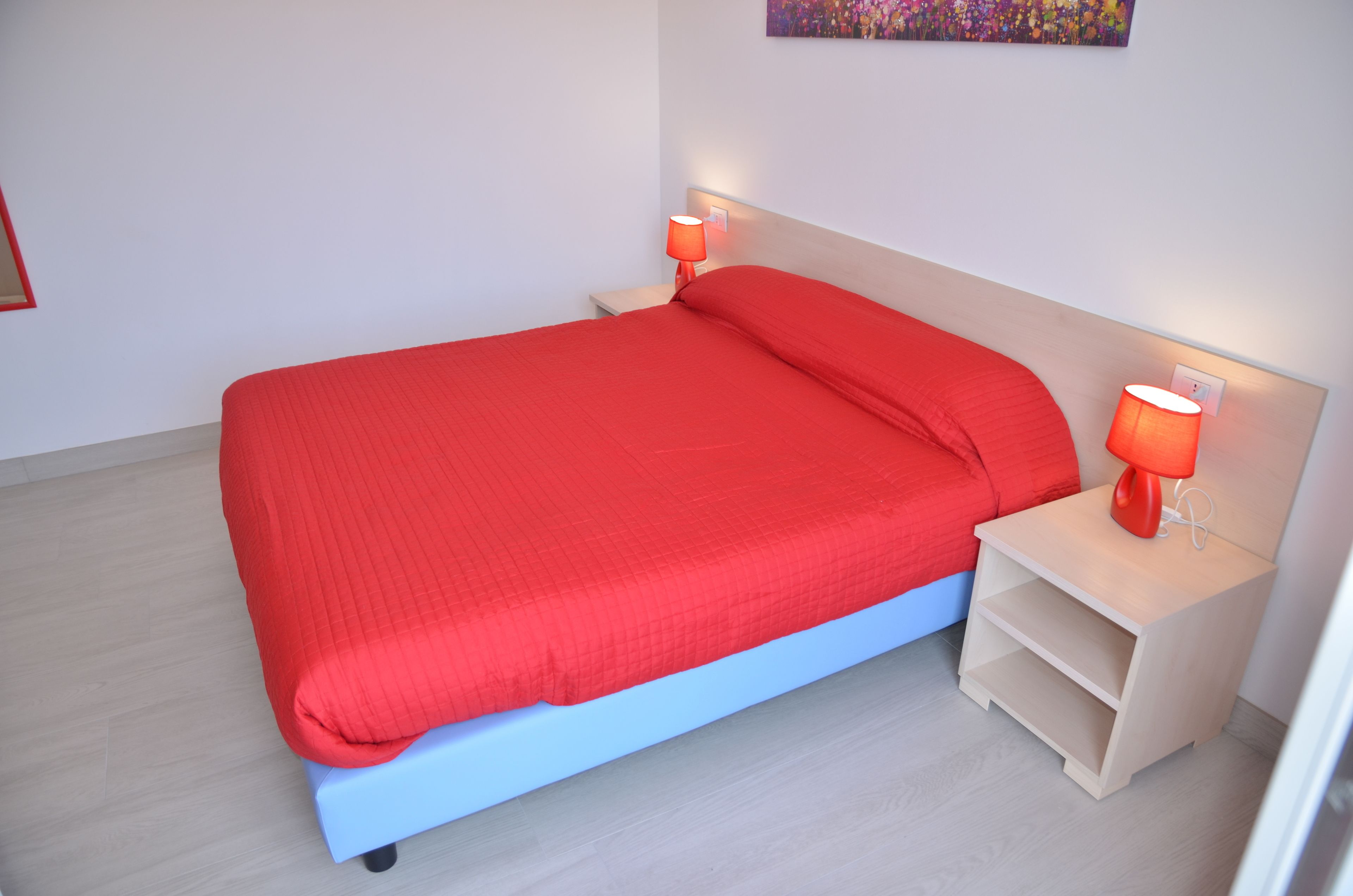 Ein Bett mit roter Bettdecke und passenden Kissen, zwei Nachttischlampen und ein heller Nachttisch mit Ablagefläche.