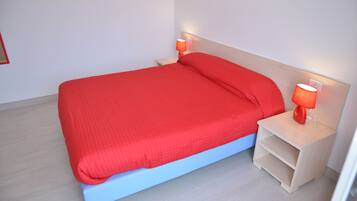 Ein Bett mit roter Bettdecke und passenden Kissen, zwei Nachttischlampen und ein heller Nachttisch mit Ablagefläche.