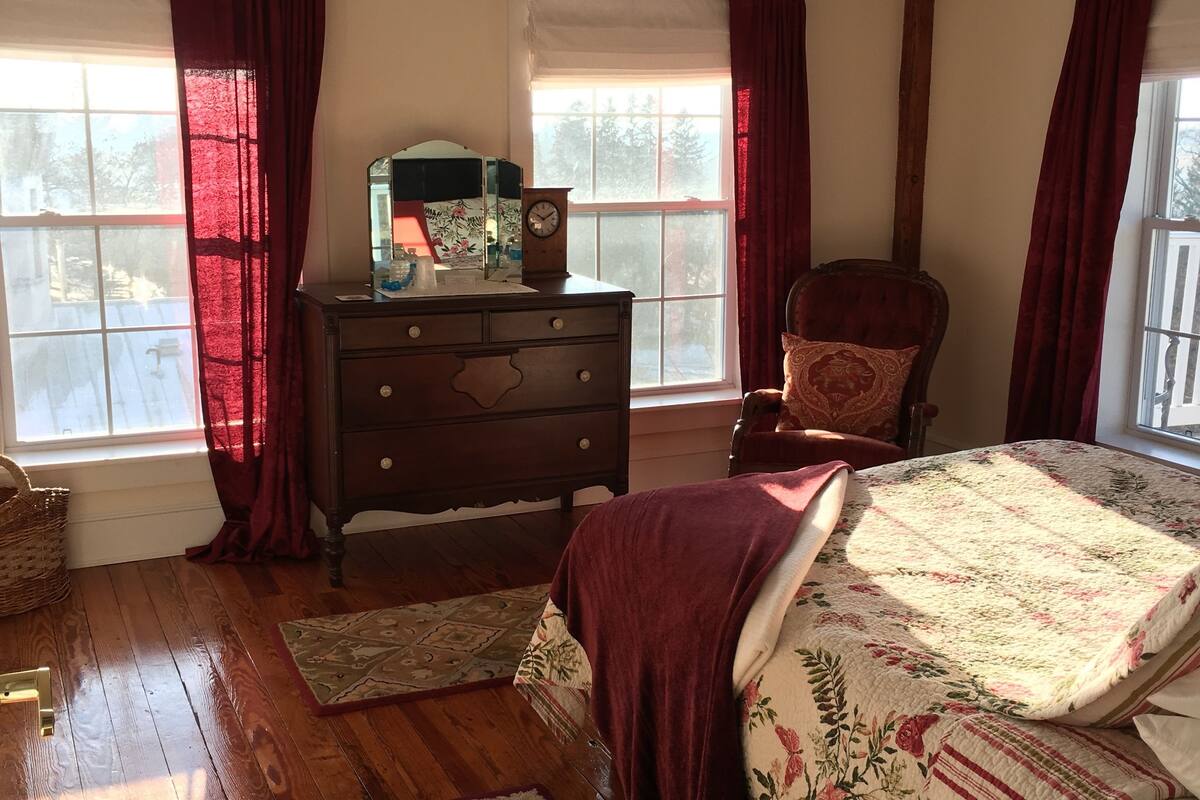 Une chambre avec un plancher en bois, un lit recouvert d’une literie à motifs floraux, une commode, une chaise et une fenêtre avec des rideaux.