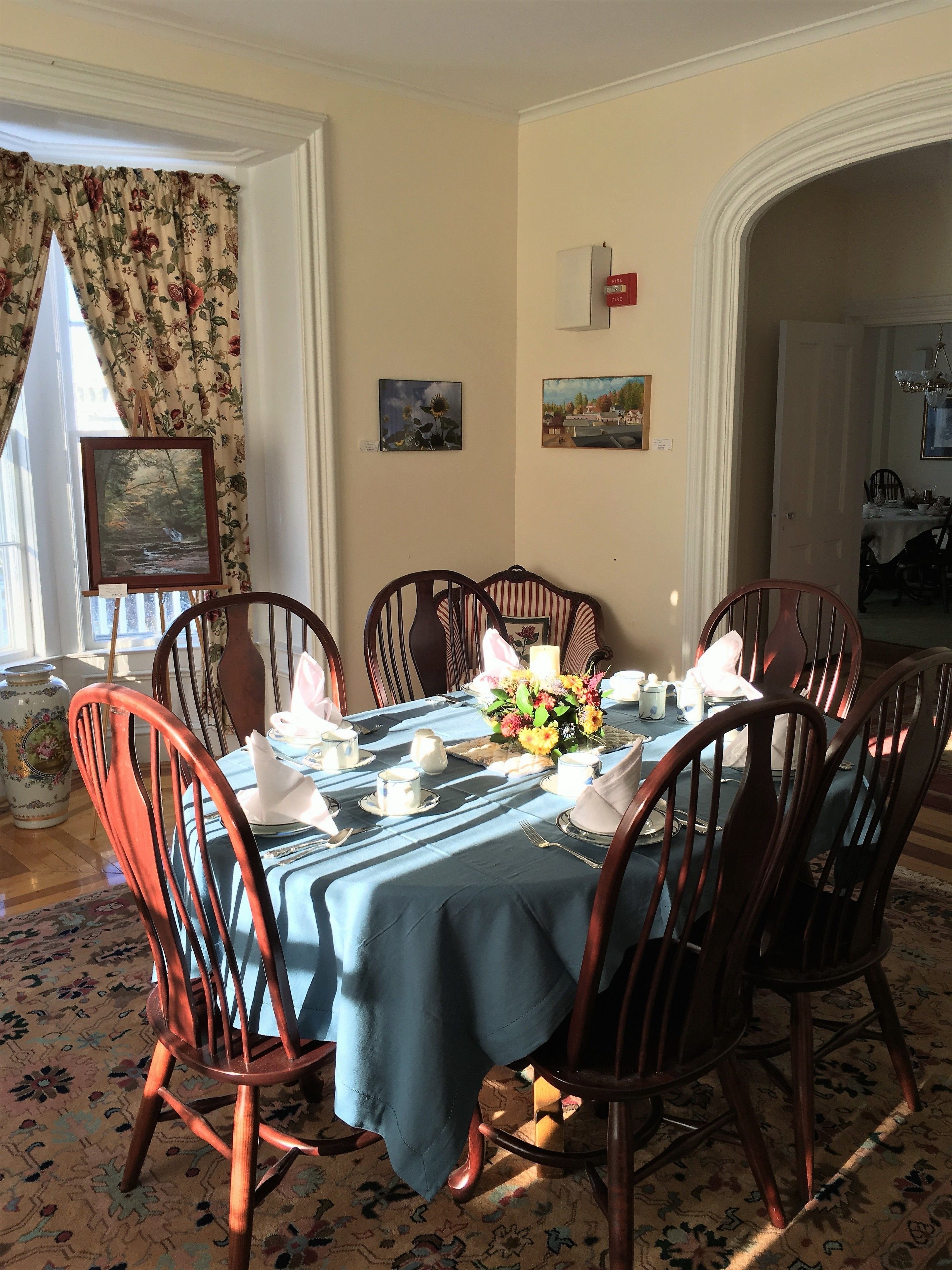 A dining room with a table set for six, surrounded by wooden chairs. There are floral curtains on the window, a painting on the wall, and a fire alarm.