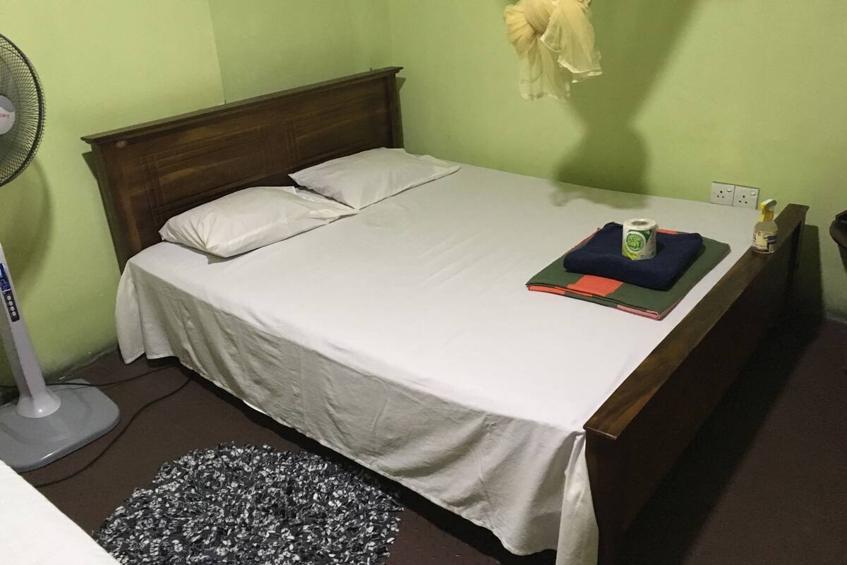 A neatly made bed with white linens, a bedside table with a bottle and a book, and a standing fan against the wall.