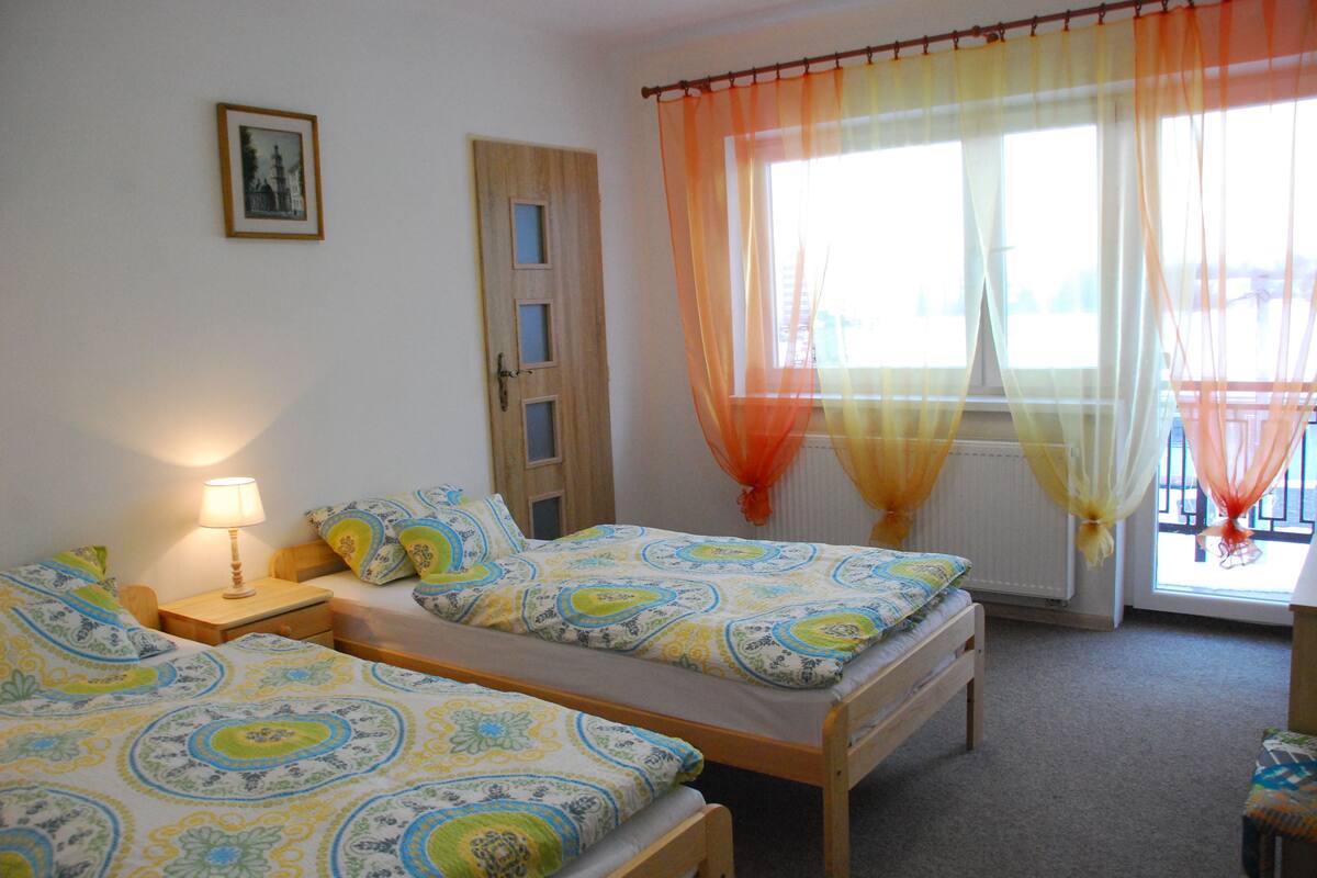 A bedroom with two single beds, a wooden door, a bedside table with a lamp, and sheer curtains over the window.