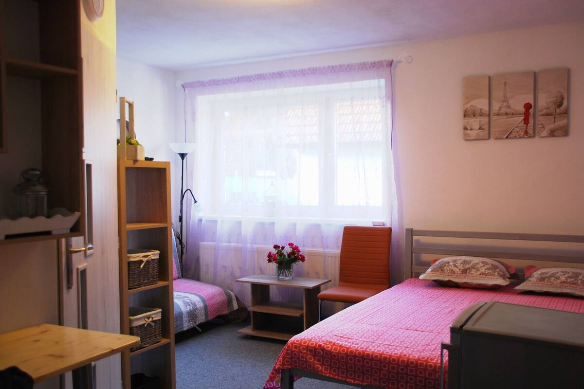 A bedroom with a single bed, a chair, a small table with a vase of flowers, and a window with sheer curtains.