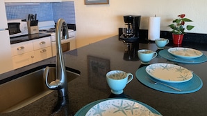 Una cocina con un lavamanos de acero inoxidable, armarios blancos, una cafetera y un cuadro en la pared.