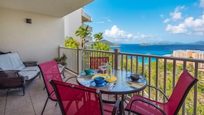 Un balcón con mesa para dos, con vista al mar y edificios, y palmeras.