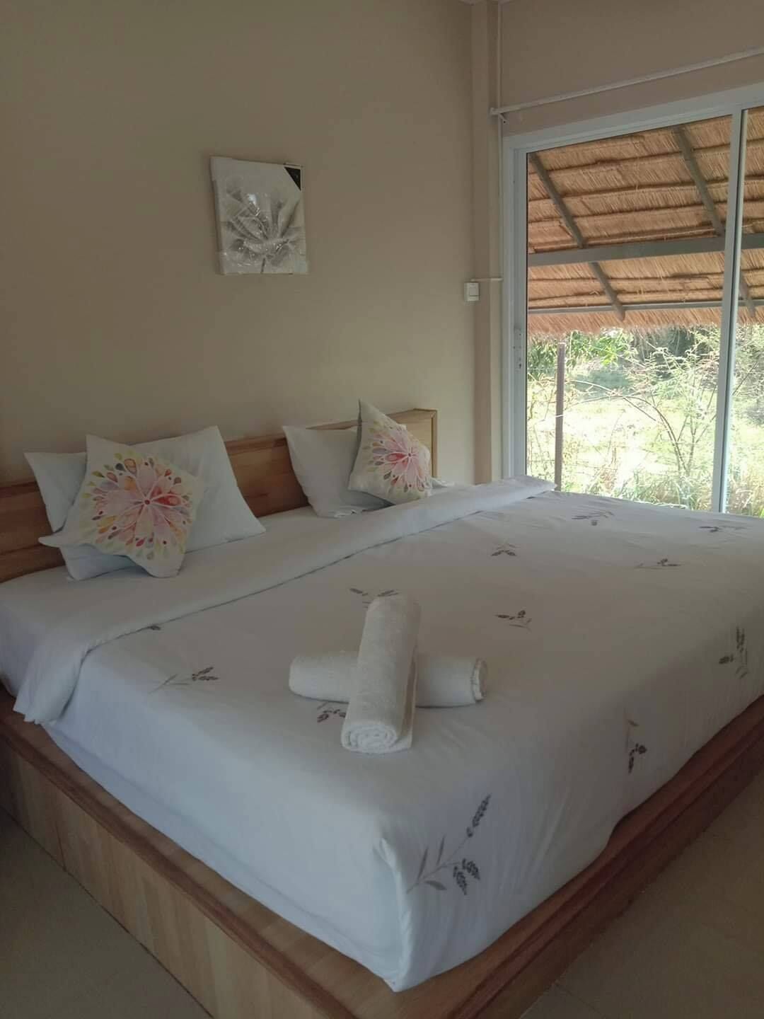 Una cama bien hecha con almohadillas estampadas, una toalla enrollada, un cabecero de madera y un ventanal con vistas a la vegetación.