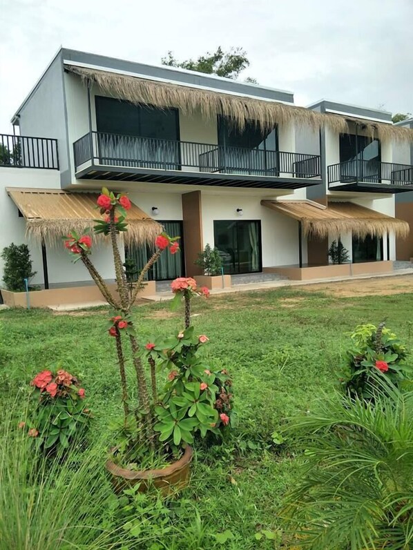 Un edificio moderno de dos pisos con balcones, techo de paja y un jardín bien cuidado con plantas en flor.