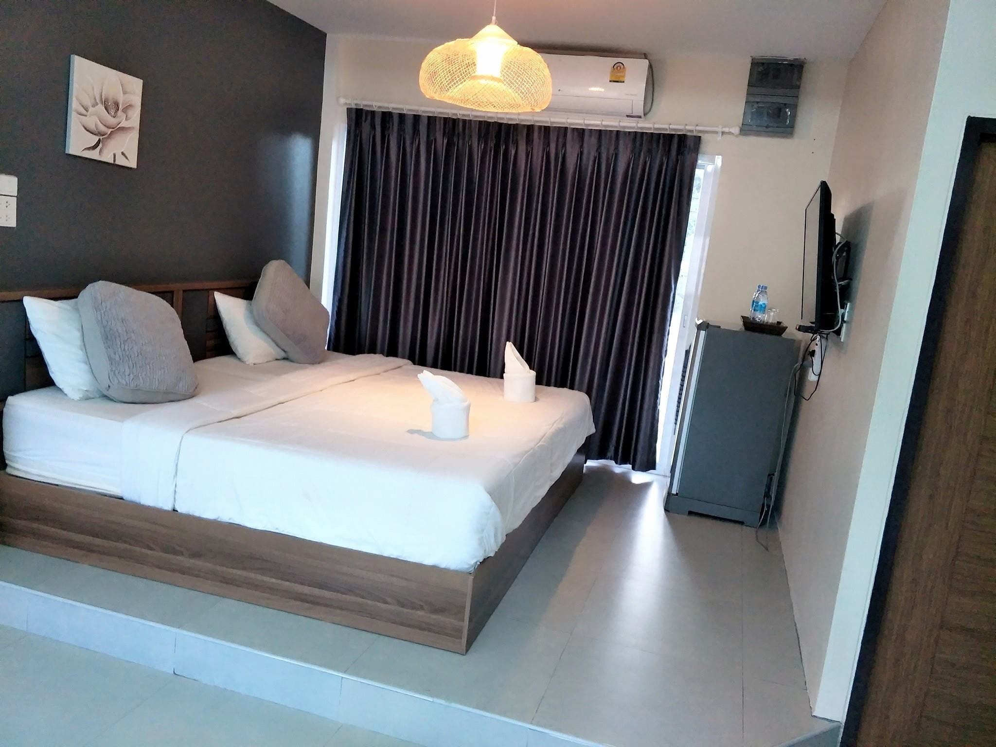 A hotel room with a bed, a bedside table with a bottle, a television, and an air conditioning unit.
