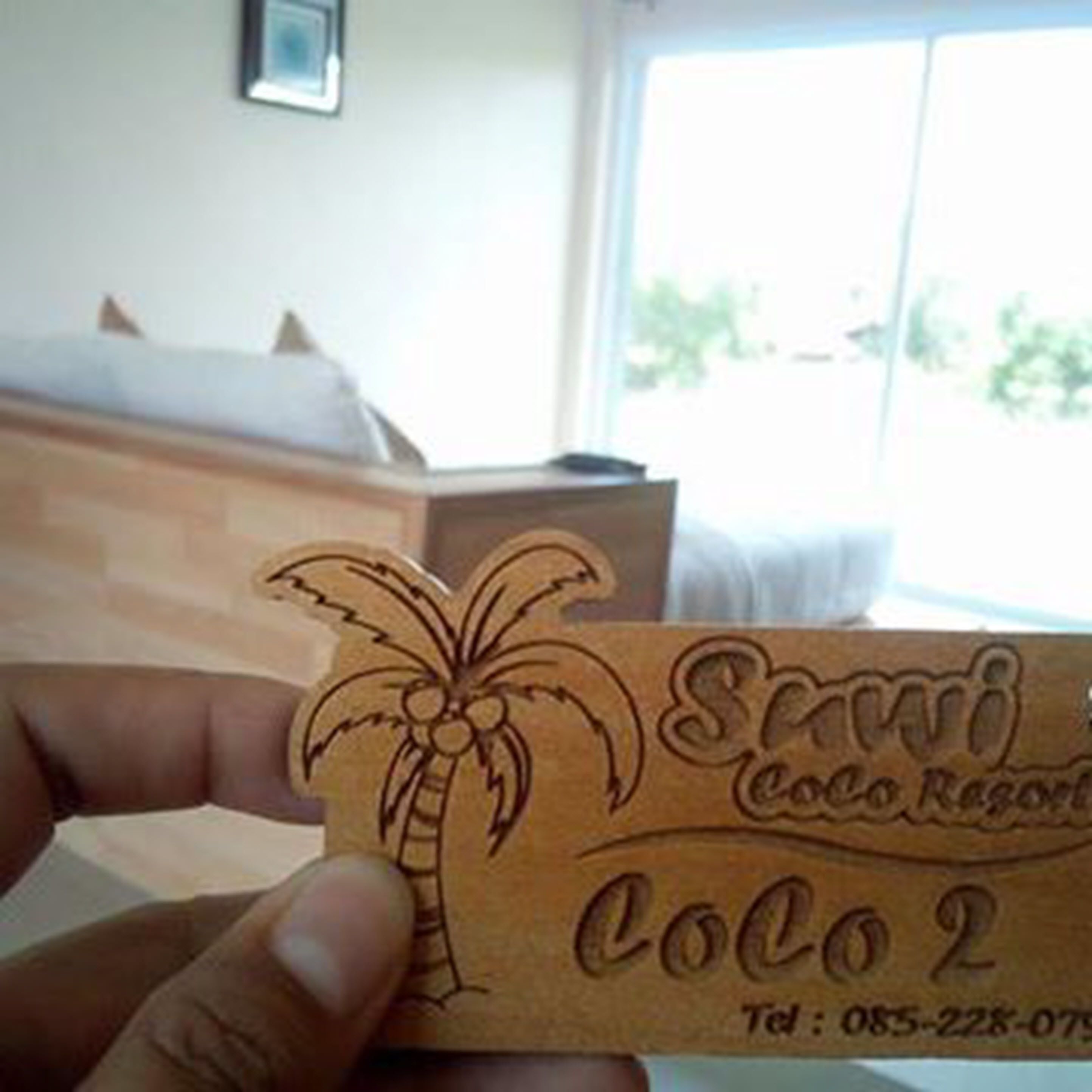 Una mano sostiene una tarjeta de madera con una palmera y texto, presumiblemente el nombre de un hotel o resort, con una vista parcial de una habitación al fondo.