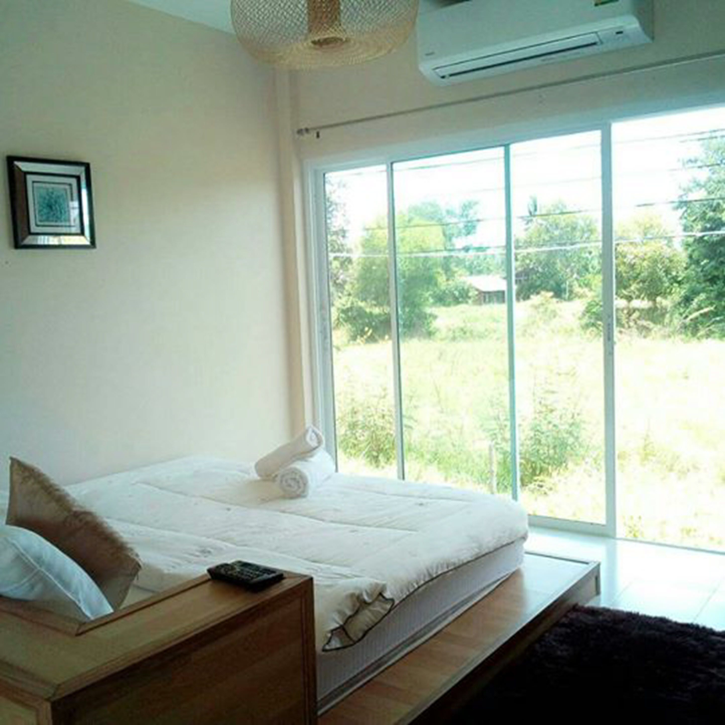 Un dormitorio con cama, mesita de noche, control remoto y una ventana grande con vistas a follaje.