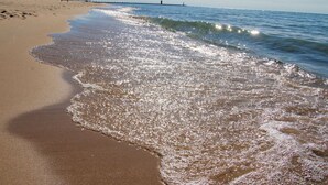 Una playa con olas suaves, cielo despejado y un muelle distante.
