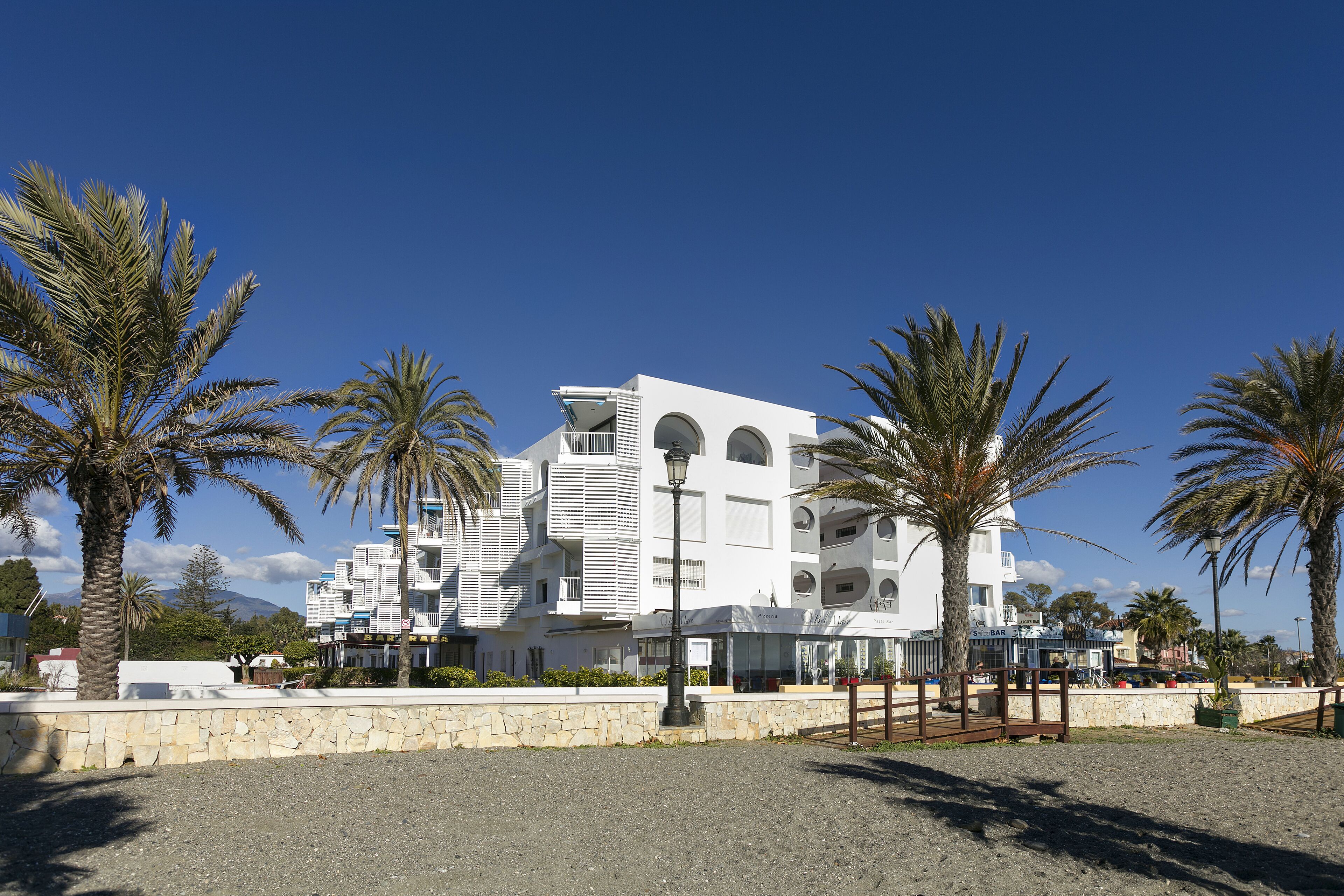 A modern white building with multiple floors, a stone wall in the foreground, and palm trees.