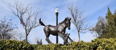 Estátua de bronze de um cavalo alado com um cavaleiro, rodeada por vegetação e um poste de iluminação.