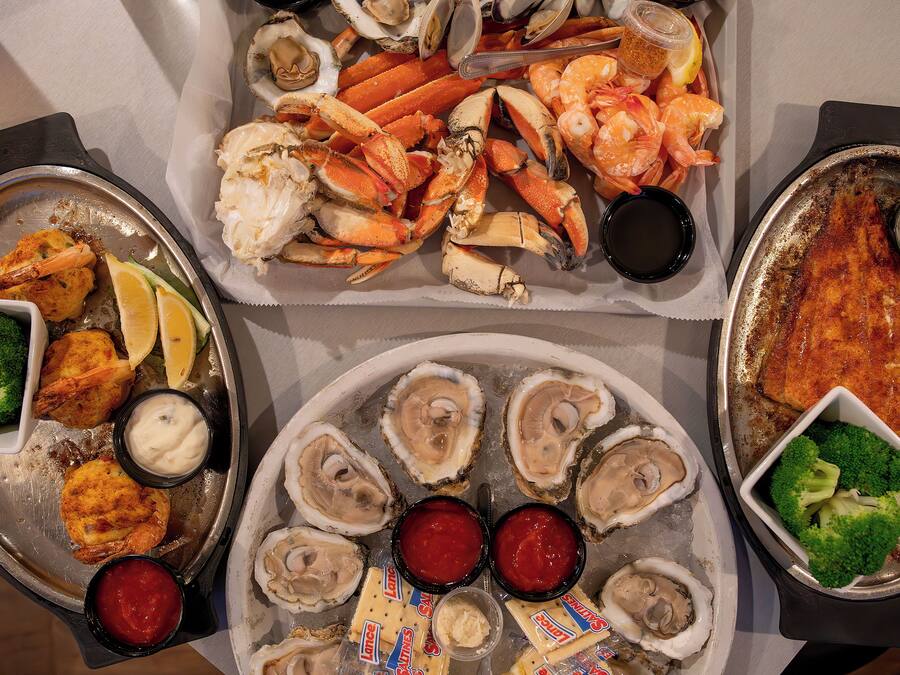 Una bandeja de mariscos con ostras, patas de cangrejo, camarones y otros productos del mar, acompañada de gajos de limón y salsas.