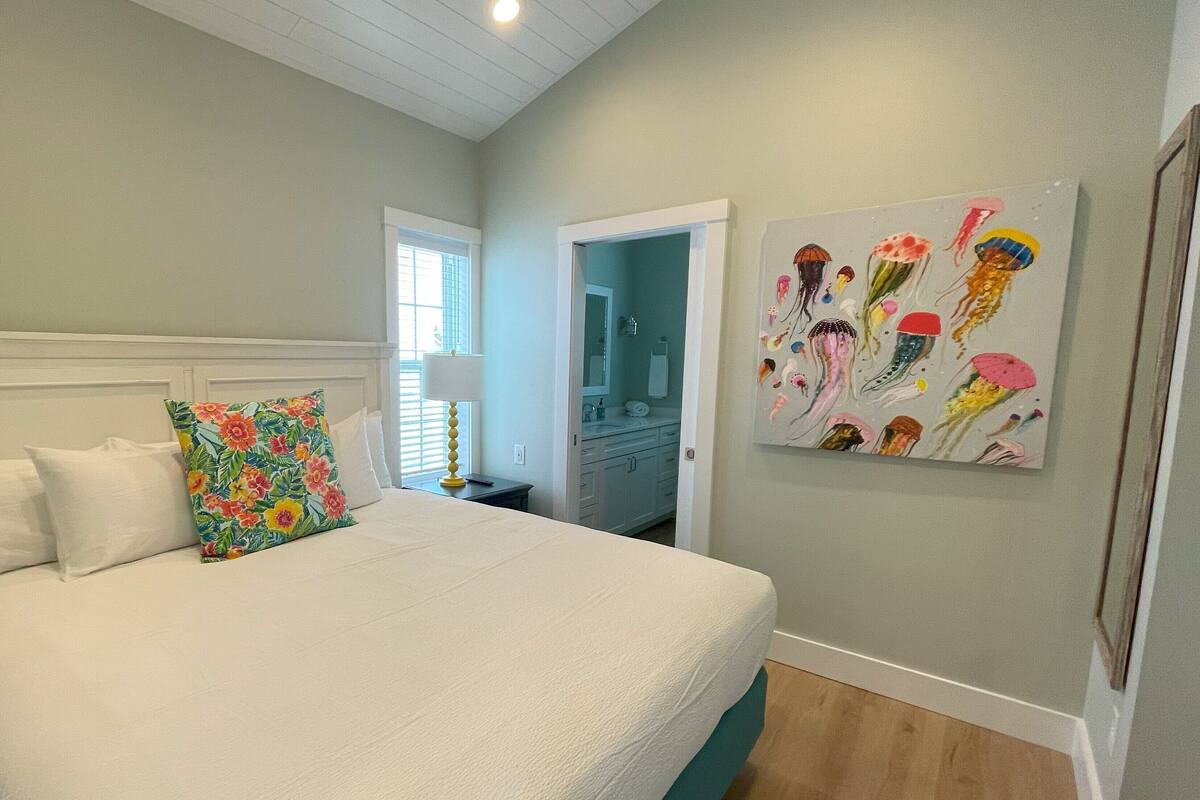 A bedroom with a bed, a window, a bathroom, and a wall with a colorful painting.