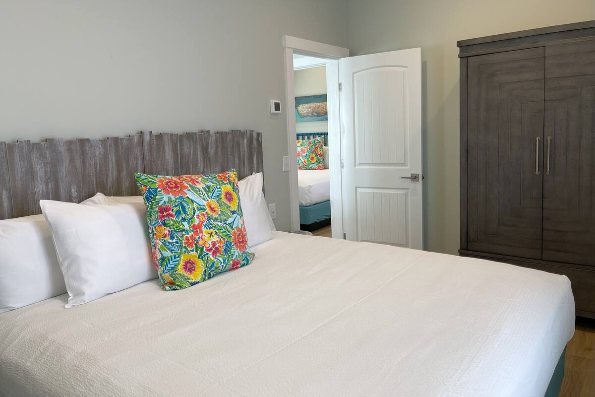 A bedroom with a bed, a colorful pillow, a wardrobe, and a door leading to another room.