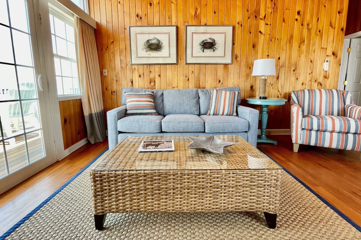 A cozy living room with a blue sofa, striped armchairs, a wicker coffee table, and wooden paneled walls.