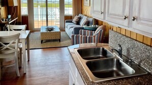 A spacious kitchen with a large window, a dining area with a table and chairs, and a living area with a sofa and cushions.