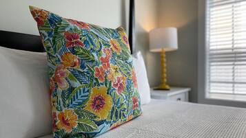 A bed with a colorful floral pillow, a white headboard, a white pillow, a gold lamp, and a window with blinds.