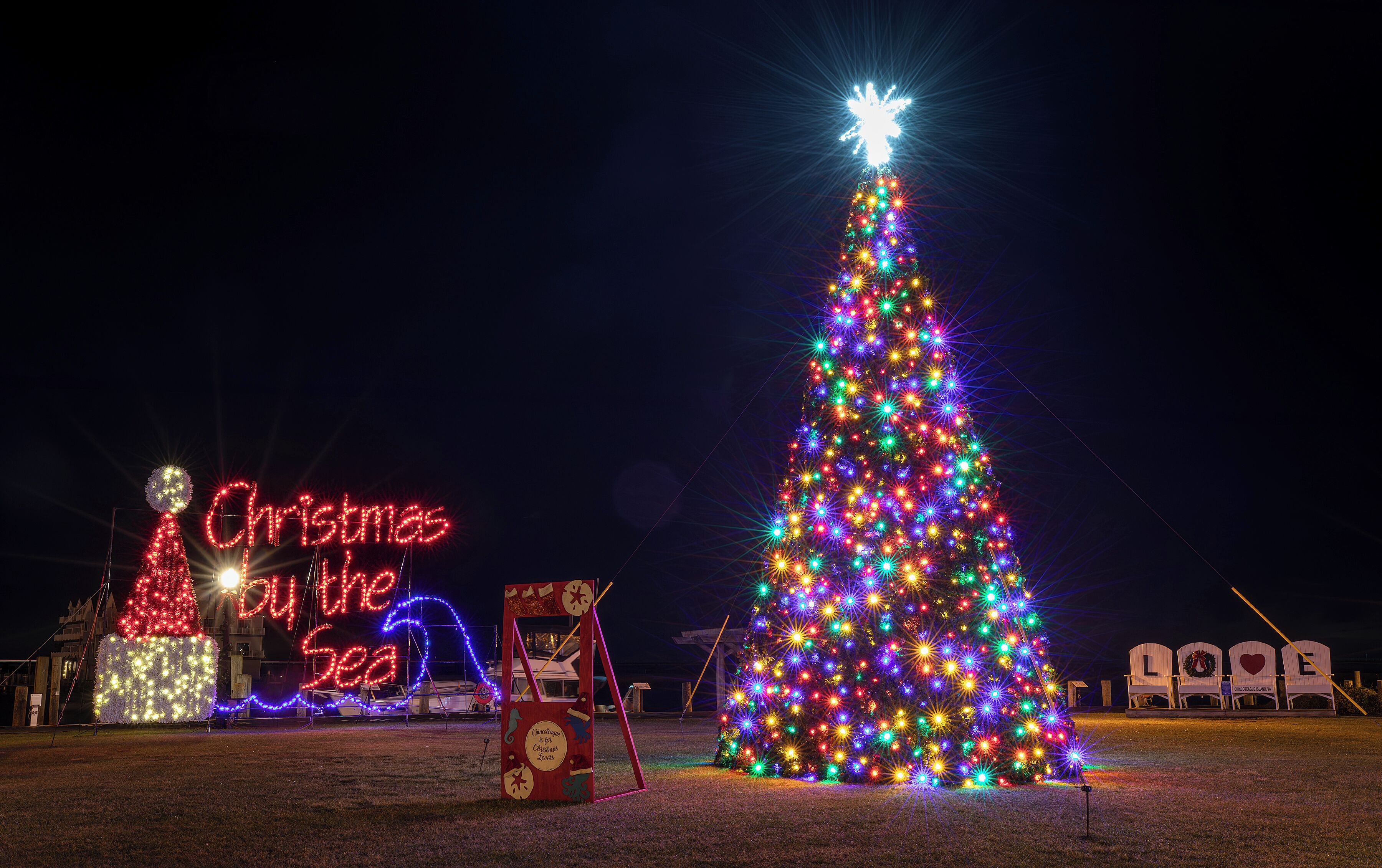 A large illuminated Christmas tree with a star on top, surrounded by various holiday decorations and a neon sign that reads "Christmas by the Sea".
