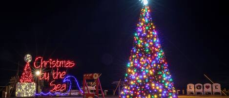 A large illuminated Christmas tree with a star on top, surrounded by various holiday decorations and a neon sign that reads "Christmas by the Sea".