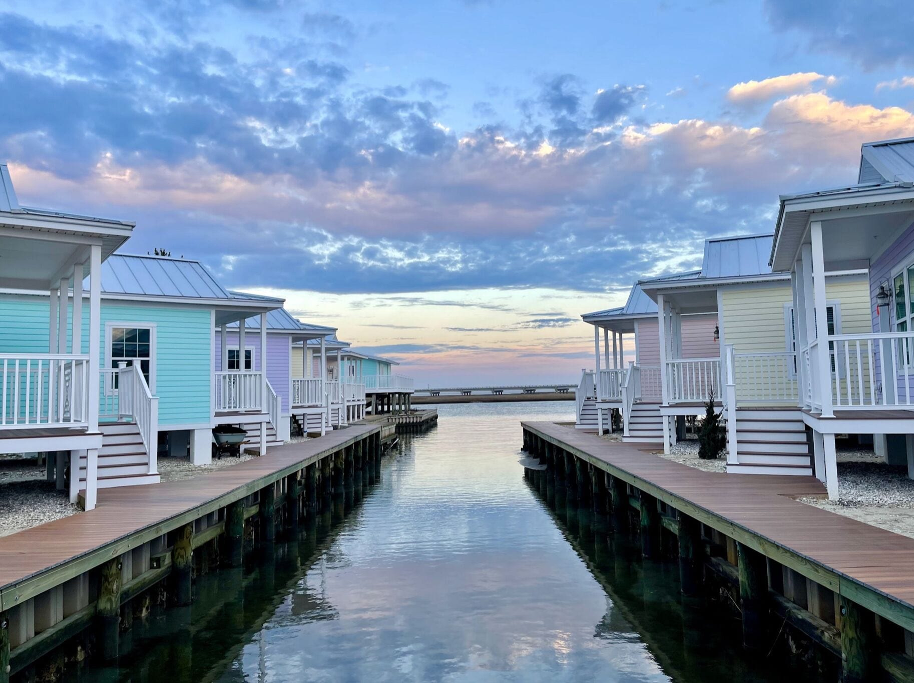A row of waterfront cabins with wooden decks and railings, connected by a dock extending into calm water.