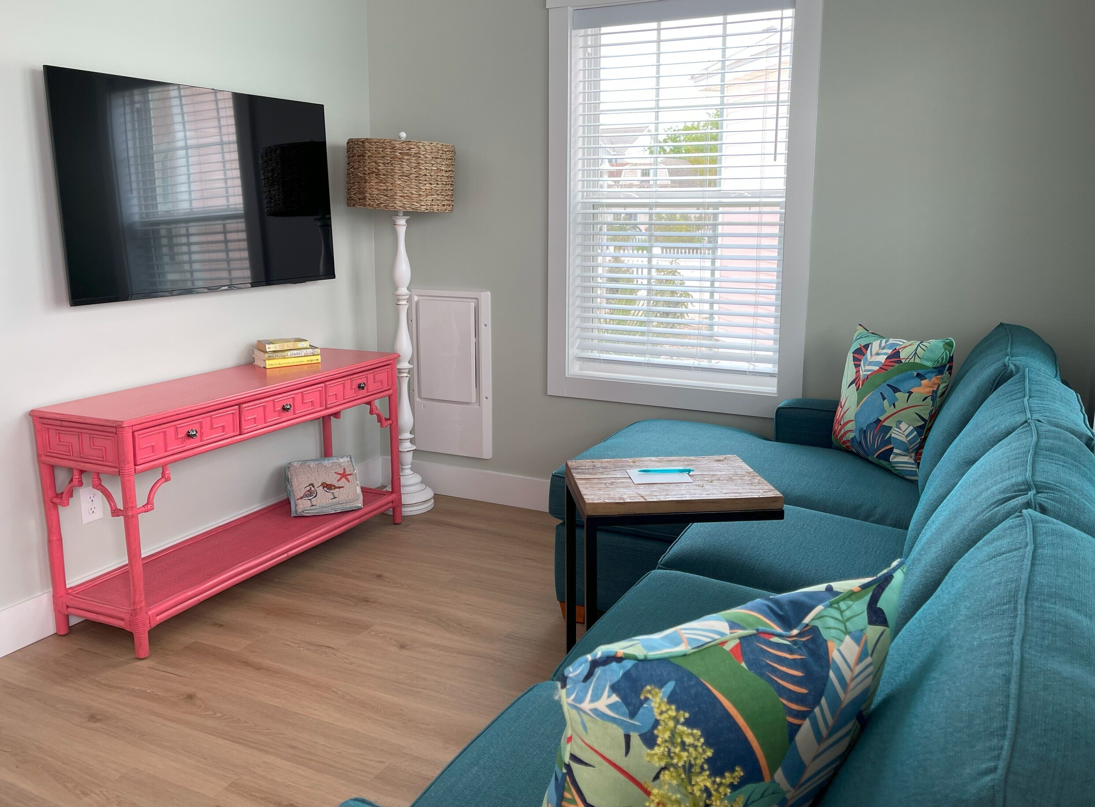 A room with a teal sofa, a pink console table, a TV mounted on the wall, and a window with blinds.