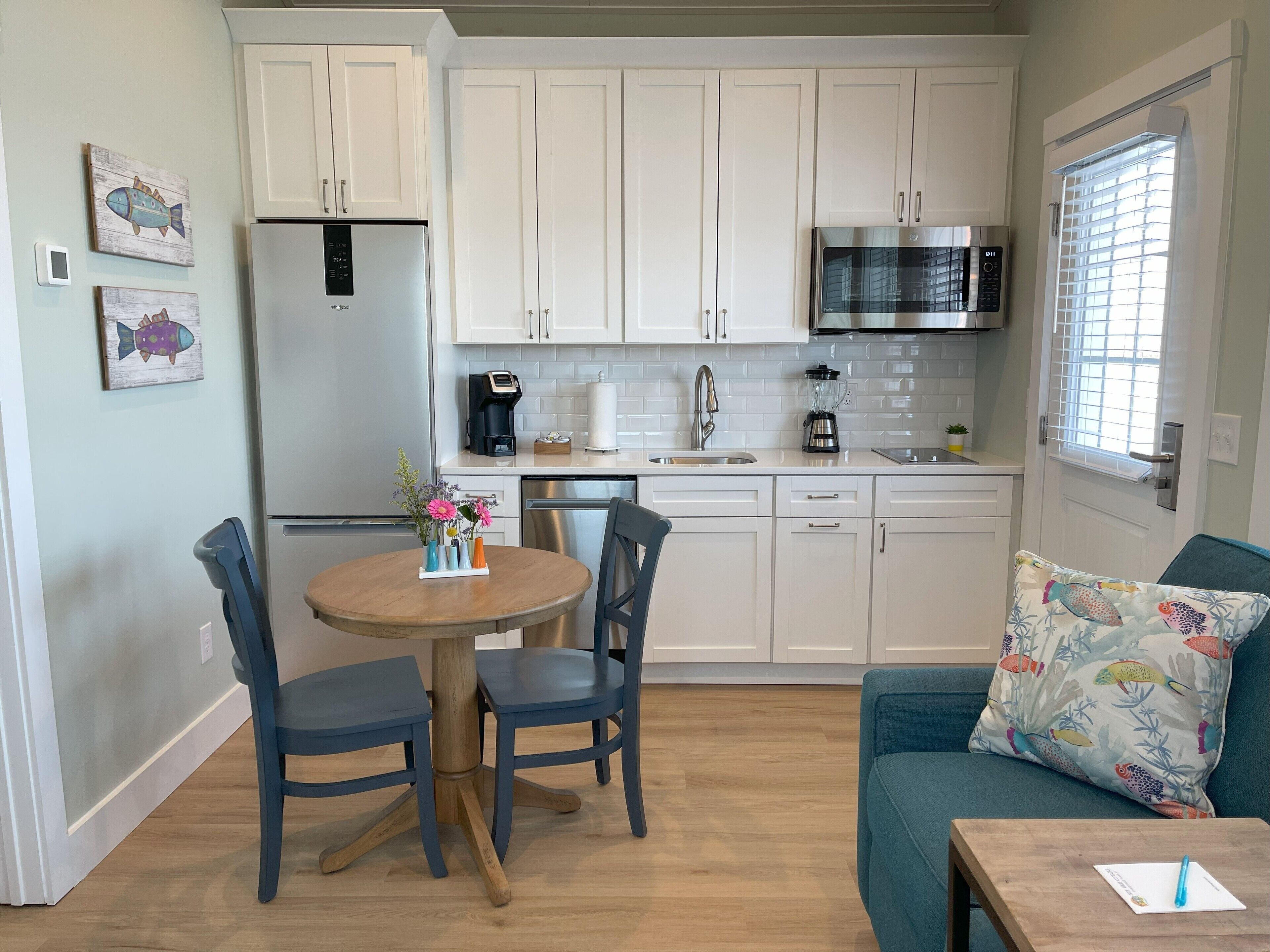 Cocina compacta con armarios blancos, microondas y cafetera. Hay una mesa de comedor con sillas y un sofá con un cojín colorido.