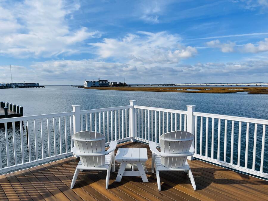 A deck with two white Adirondack chairs and a small table, overlooking a calm body of water with a distant shoreline and a pier.