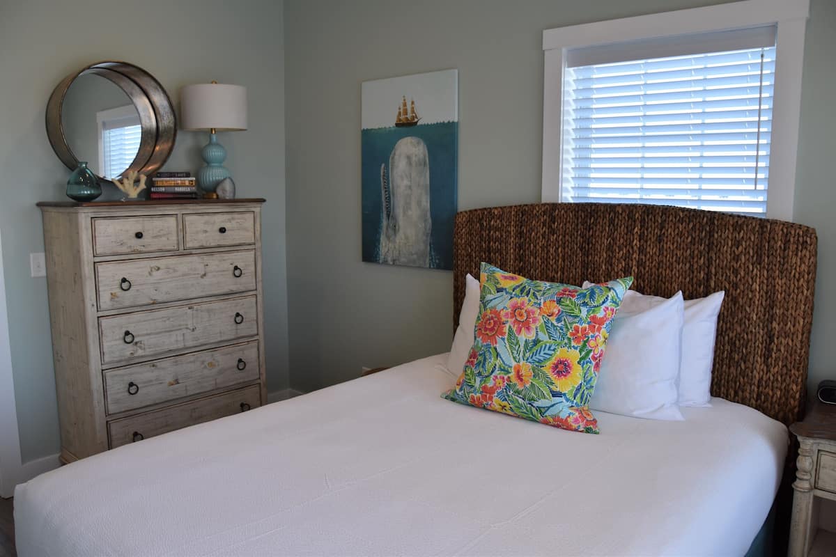 A bedroom with a bed, a dresser, a mirror, a lamp, and a painting of a ship and whale.