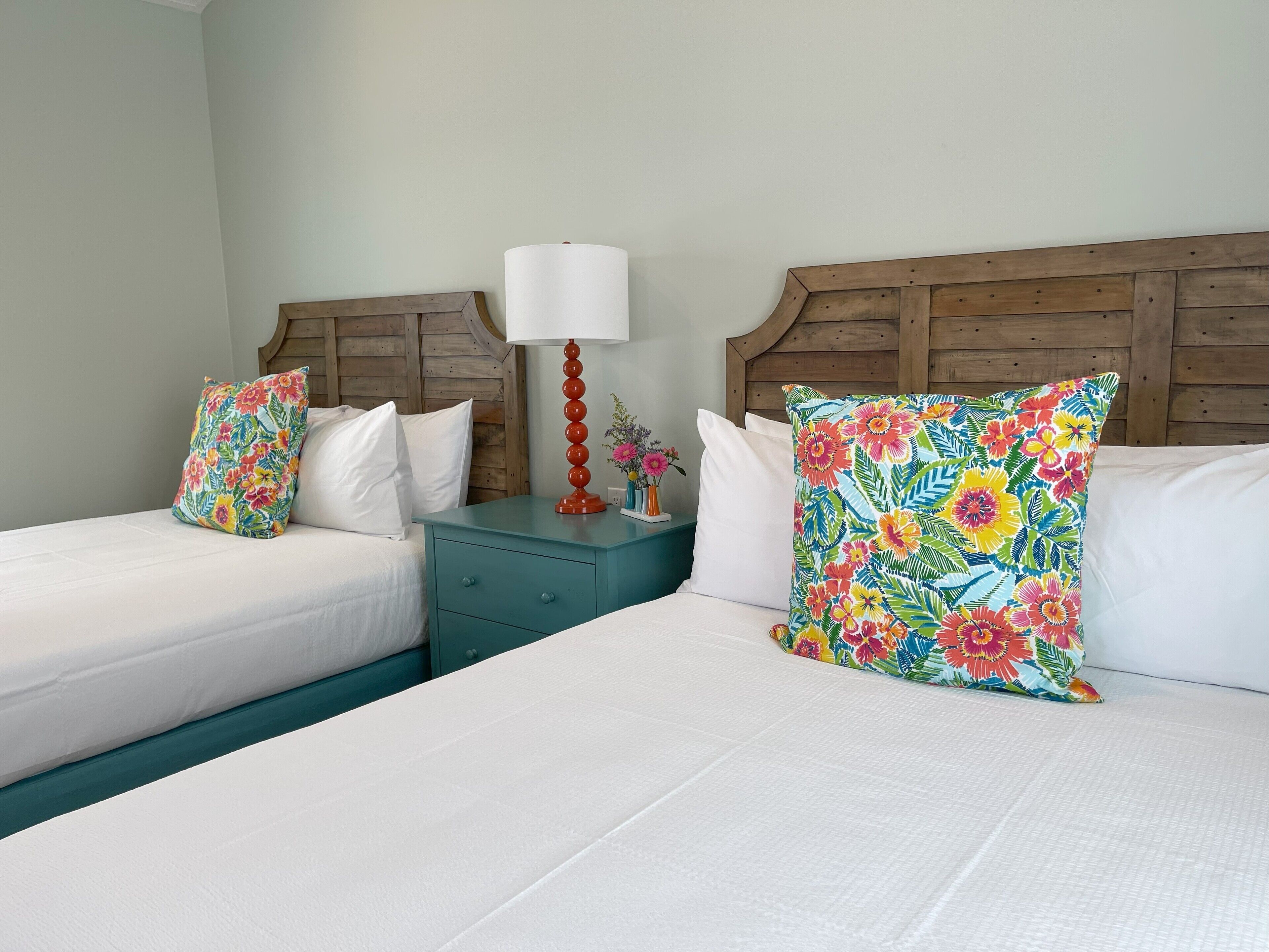 Habitación de hotel con dos camas, cada una con ropa blanca y almohadillas florales de colores. Hay una mesita de noche color turquesa con una lámpara roja y un florero con flores.