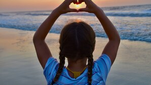 A person making a heart shape with their hands against a sunset at the beach.