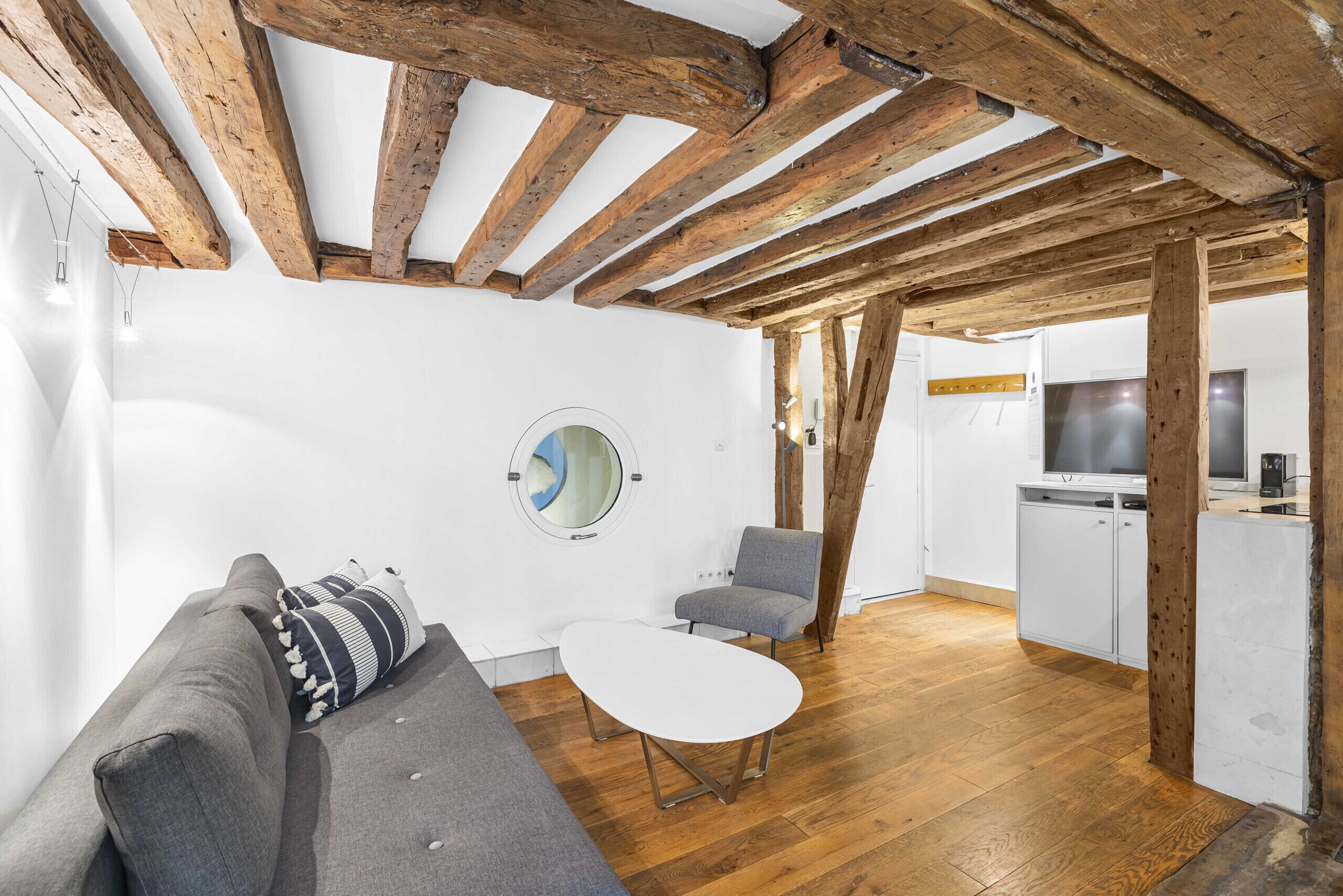 Een moderne woonkamer met een houten plafond, een ronde spiegel, een grijze bank, een witte salontafel en een grijze fauteuil.