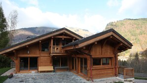 Eine Holzhütte mit einer Steinterrasse, großen Fenstern und einem Balkon mit Blick auf eine Berglandschaft.