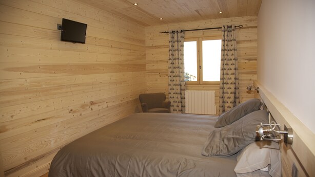 Een kamer met houten lambrisering, een bed, een stoel, een raam met gordijnen met een patroon en een wandtelevisie.