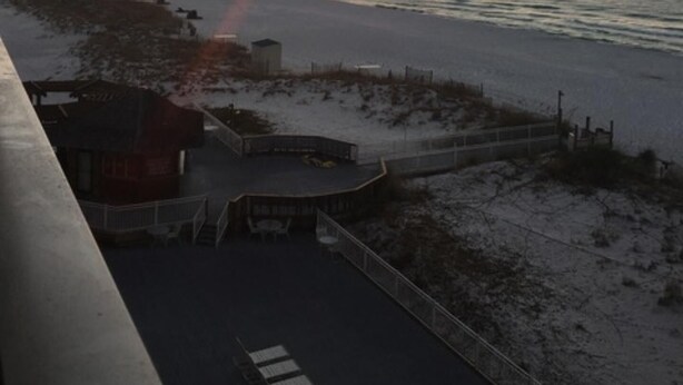 Une vue sur le coucher de soleil depuis un balcon donnant sur une plage, avec un petit bâtiment et une zone délimitée par une clôture.