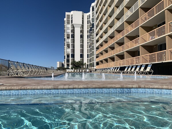 Une piscine à l’eau claire, entourée de chaises longues et bordée d’un bâtiment doté de nombreux balcons.