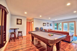 Una mesa de billar en una habitación con suelo de madera, un sofá y una puerta que da a otra estancia.