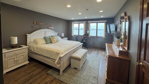 Un dormitorio con cama, mesitas de noche, una banca, cómoda, televisor y ventanas con vistas a una masa de agua.