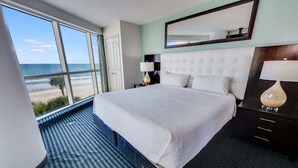 A hotel room with a large bed, a nightstand, a lamp, a mirror, and a view of the beach.