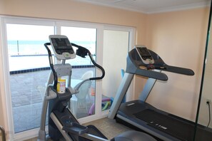 A home gym with two treadmills and a stationary bike, a window with a view of the beach, and a door leading to a patio area.