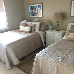 Een slaapkamer met twee bedden, een nachtkastje met een lamp, een lijst met een afbeelding aan de muur en een decoratief kussen op een van de bedden.