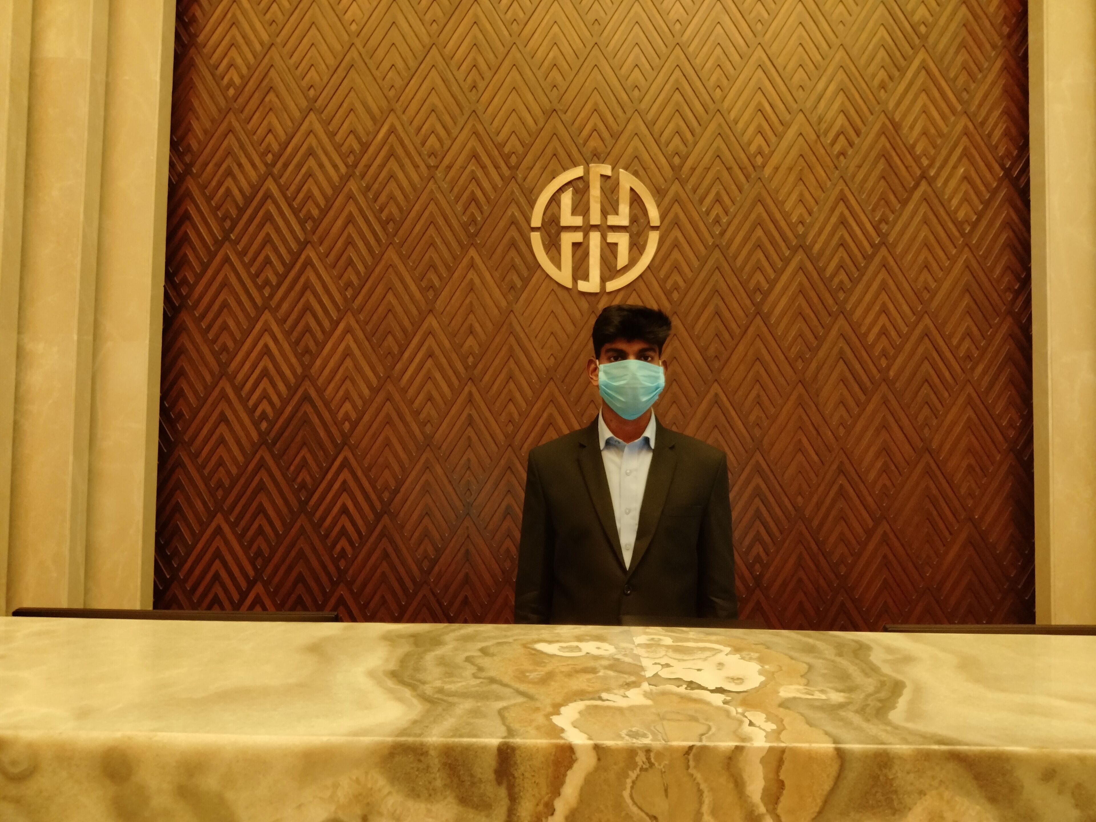 A man in a suit standing at a reception desk with a patterned wall behind him.