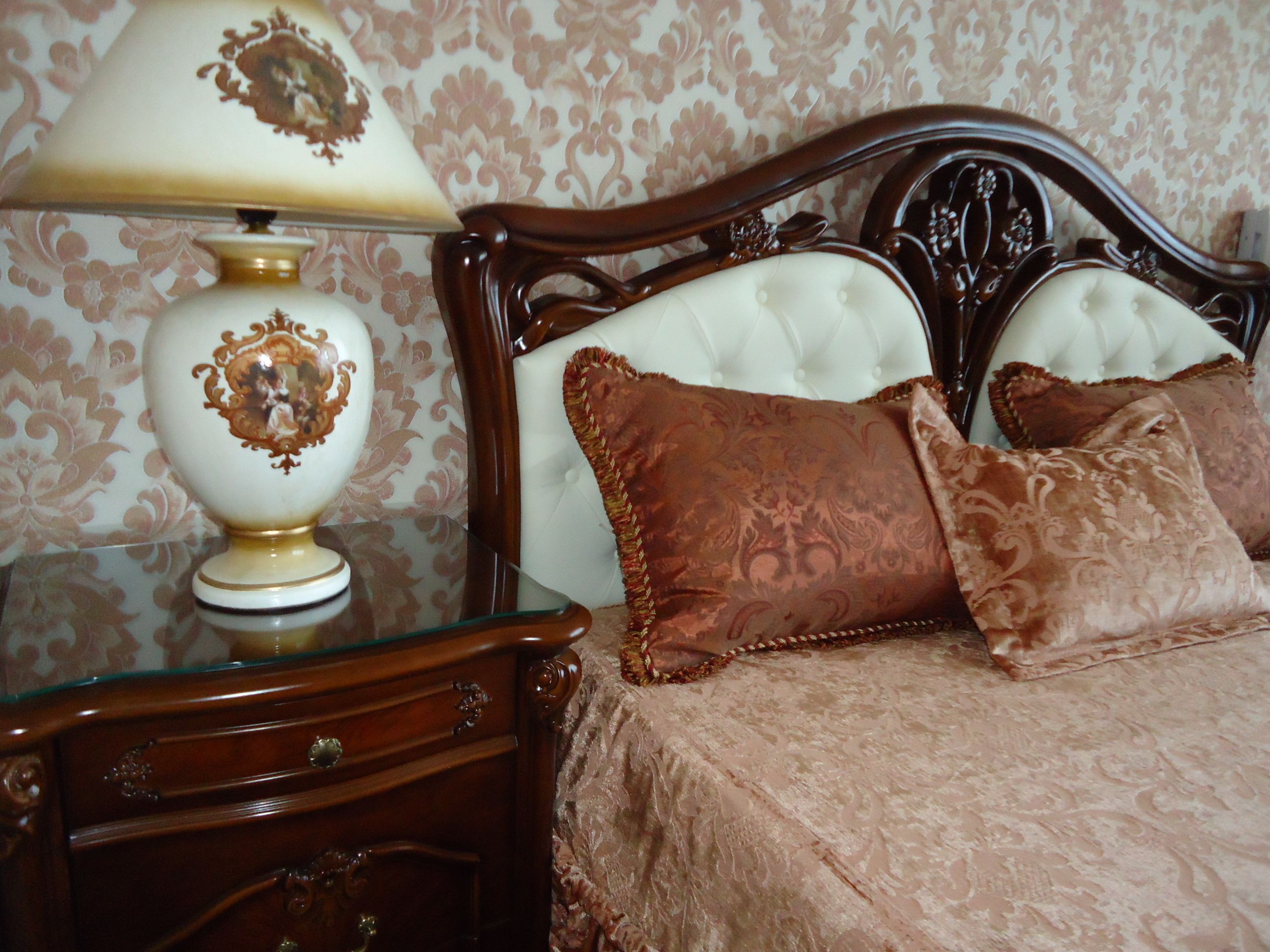 A bedroom with a wooden headboard, a glass bedside table with a lamp, and a bed with a patterned bedspread and pillows.
