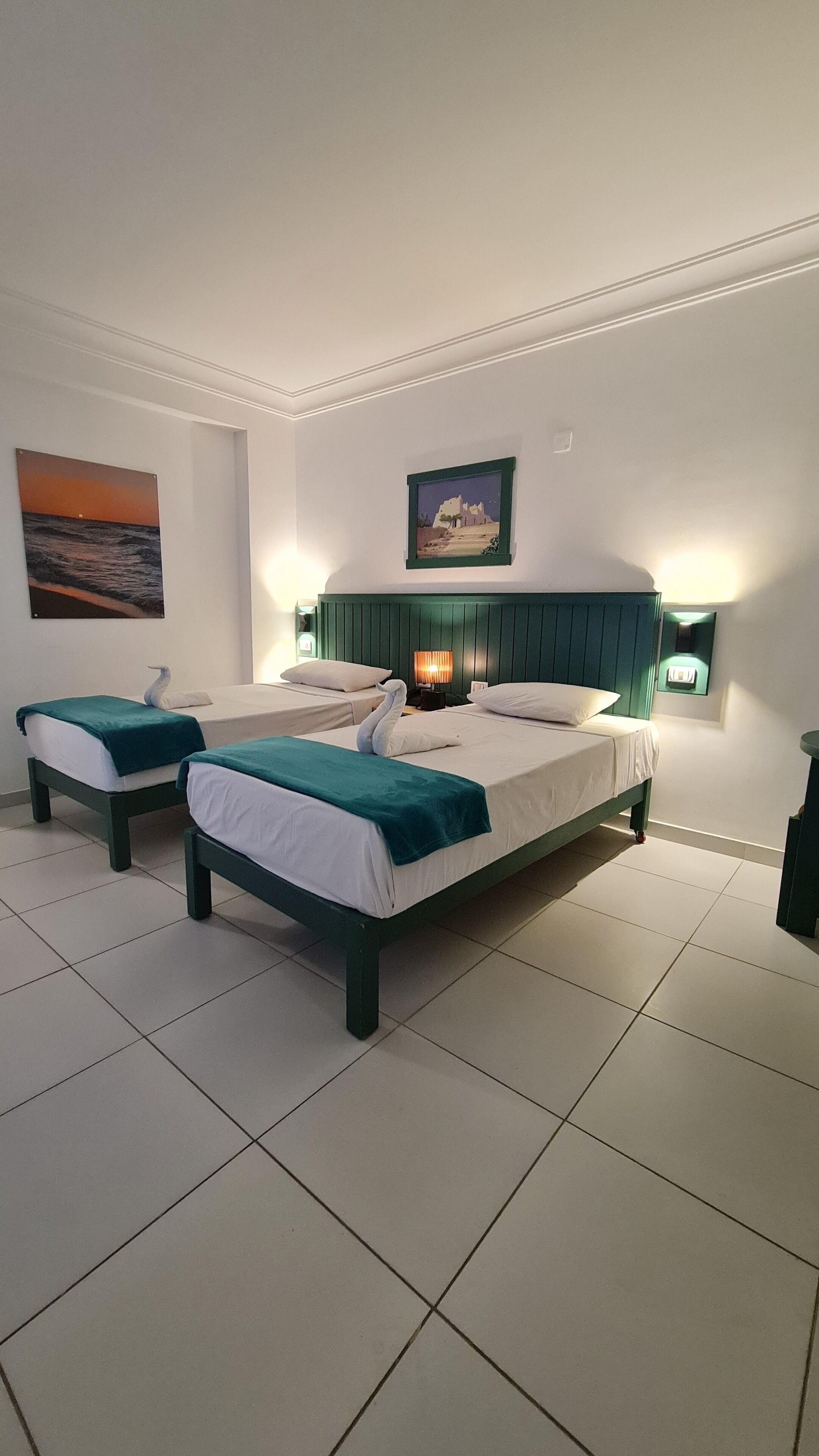 A hotel room with two single beds, a large bed, a nightstand with a lamp, a painting on the wall, and a view of the sea.