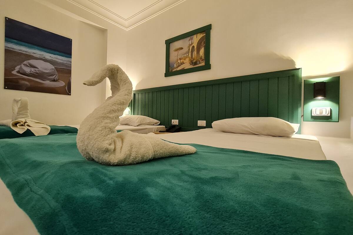 A hotel room with two beds, each with a green bedspread and white pillows. There are wall-mounted paintings, a green headboard, and a wall-mounted light fixture.