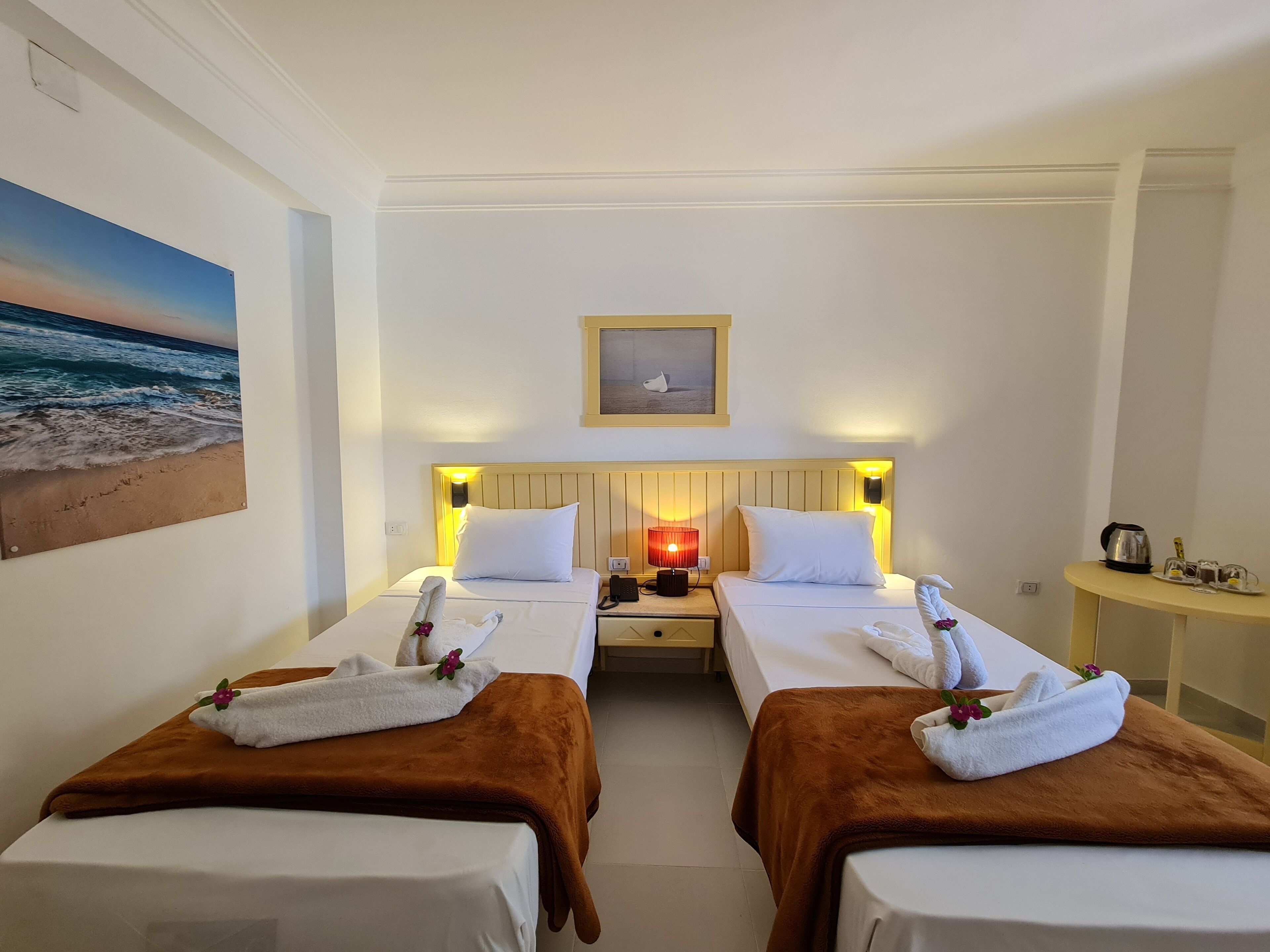 Ein Hotelzimmer mit zwei Betten, einem Nachttisch mit Lampe, einem kleinen Tisch mit Wasserkocher und Gläsern sowie einem Bild einer Strandansicht an der Wand.